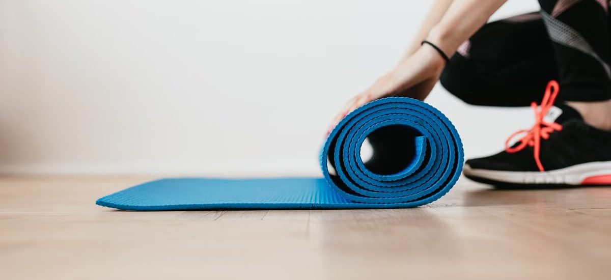Side view of unrecognizable female athlete in black leggings and sneakers unfolding sport mat for fitness training indoors